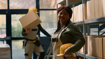 Wall Mural - Portrait of cheerful african american warehouse sorter in protective uniform dealing with packaged stock in cardboard boxes ready to be shipped, happy to work in professional distribution center