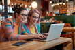 Coworking. Work on line. Two young ladies in colorful clothes, happily working on a laptop at a cafe table. Soft, cinematic light. Senior adult woman with glasses using laptop.