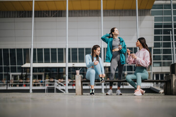 Wall Mural - Three young women in sportswear taking a break and enjoying snacks outdoor, embodying an active city life.