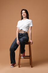 Poster - Smiling tattooed woman sitting on stool against beige background