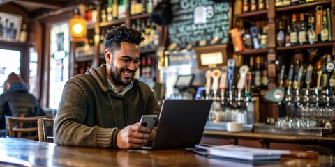 Contented male utilizing cellphone while working on laptop at a bar.