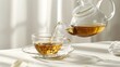 A clear glass teapot pouring green tea into a matching cup on a white background