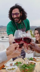 Wall Mural - Vertical HD video of young group of diverse people toasting red wine at summer party celebration. Multiracial friends sitting around dinner table while enjoying bbq dinner together at terrace.