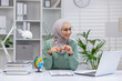 © Liubomir - A cheerful Muslim woman wearing a hijab engaged in online teaching, using a laptop and pointing at a small globe, surrounded by books in a well-organized office.