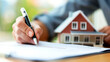 A person is signing a document with a pen. There is a model house on the table. The background is blurred.