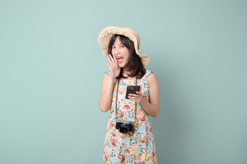 Wall Mural - Travel and vacation promotion concept. Happy Asian woman wearing casual dress and hat with camera and mobile phone while shouting to free copy space isolated on pastel green background.