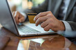© Nalinthip - Close-up of a businessman sitting on a table holding a credit card and using a laptop to record data.