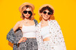 Two young beautiful smiling brunette hipster female in trendy summer costume clothes. Sexy women posing near yellow wall in studio. Positive models having fun. Cheerful and happy. In hat, sunglasses