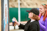 Fototapeta  - Two female artists painting picture with paint spray can spraying it onto canvas at outdoor street exhibition, side view of passionate female art makers