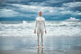 Fototapeta  - Happy hairless girl with alopecia in white futuristic suit stands on beach bathed by ocean waves, metaphoric performance of bald strong female artist, overcoming challenges of life and self confidence