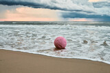 Fototapeta  - Large pink sphere lapped by waves against seashore against horizon and storm clouds, pink ball symbol of serenity contrasts against brewing turbulence in sky and relentless energy of sea