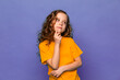 Thoughtful little girl in orange t-shirt looks away and touches her mouth with her finger. Emotions and reflections, lilac background