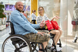 Fototapeta Młodzieżowe - Smiling man in a wheelchair and woman with shopping bags enjoying time at the mall