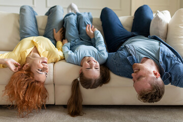 Upside down portrait of joyful family lying on sofa with heads down, mother, father and daughter having fun at home