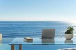 A simple and serene writing desk with a clear glass top, a minimalist laptop, and a tranquil sea view outside