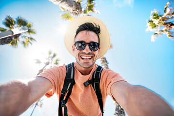 Wall Mural - Happy tourist taking self portrait outside with cellphone on summer vacation - Handsome young man laughing at camera enjoying summertime day out - Tourism, traveler life style and technology concept