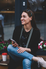 Wall Mural - Trendy positive female student sitting on bench with mobile phone in hands while notepad and takeaway coffee staying near, happy woman smiling and looking away waiting update notification on cellphone