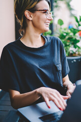 Cheerful hipster girl looking aside and smiling in break of course language online via laptop, positive female freelancer casual dressed enjoying time with netbook at street cafe while waiting friend