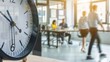© photolas - A clock displays the time with motion-blurred individuals bustling in the office background, suggesting a hectic workday atmosphere.