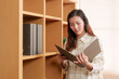 © Ekkasit A Siam - Young Asian woman in plaid shirt reading folder in office setting. Wooden bookshelf with files in background. Scene portrays focused office environment, professional atmosphere.
