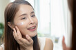 © Pormezz - Facial beauty skin health care, smile of cute asian young woman looking at mirror, hand applying moisturizer lotion on her face, female putting cream treatment before makeup cosmetic routine at home.