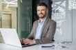 © Liubomir - Cheerful businessman smiling while working on his laptop in a modern office setting. Professional work environment with a touch of technology and innovation.