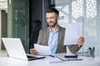 © Liubomir - Smiling businessman reviewing documents while working on a laptop in a modern office. Professional office setting with notepad and penholder in the background.