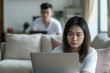 Young adult woman focusing intently on her laptop screen in a brightly lit living room, with a blurred male figure in the background, symbolizing work from home or remote learning
