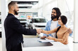 © Prostock-studio - A man and a woman are shaking hands inside a well-lit car showroom. The man appears to be a car salesman, and Indian woman is a potential customer. They are standing next to a shiny car