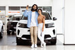 © Prostock-studio - Indian man and woman are standing in front of a car, looking towards the camera. The car is parked on a paved road with trees in the background.