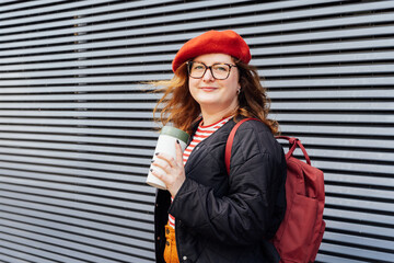 Young smiling fashion plus size woman with reusable coffee cup on striped wall background. Body positive. Female freedom. Urban street fashion style. Confident happy satisfied overweight model