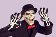 Man in Halloween outfit scares and screams making scare gesture on lilac background in studio. Close up of young man with skull painted on face and wearing suit and hat raises his arms and scares you.