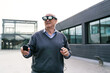 © Koldo_Studio - Senior businessman engaging with virtual reality glasses and a controller, interacting with virtual elements outside a modern office building
