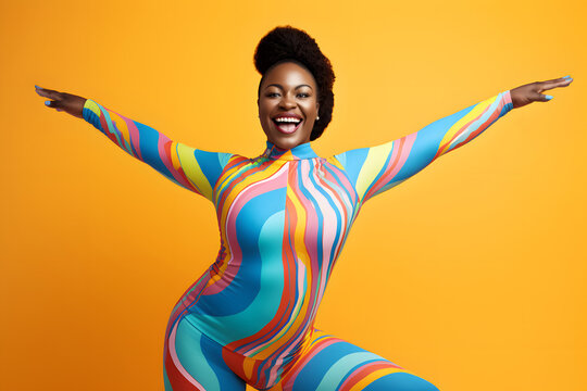 Body positivity: joyful curvy black woman with afro hair wearing a multicolor one-piece yoga jumpsuit is dancing with outstretched arms against a plain yellow background and smiling.