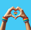 Hands forming heart shape against bright blue sky, rainbow colored wristbands, symbolizing LGBTQ