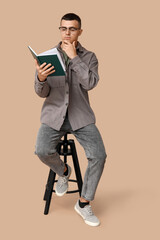 Poster - Pensive young man sitting on chair and reading book on beige background