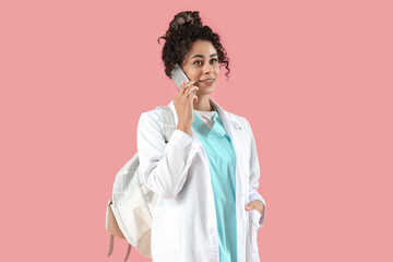 Poster - Female African-American medical intern with backpack talking by mobile phone on pink background