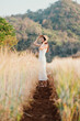 © Satori Studio - A woman in a white dress stands in a field of tall grass. She is wearing a straw hat and looking up at the sky. The scene is peaceful and serene