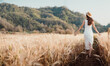 © Satori Studio - A woman is walking through a field of tall golden grass. She is wearing a white dress and a straw hat. The scene is peaceful and serene, with the woman enjoying the beauty of nature
