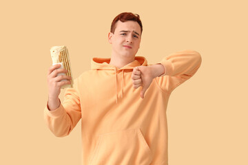 Wall Mural - Young man holding tasty doner kebab and showing thumb-down on beige background