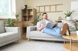 © Pixel-Shot - Young man with bottle of cucumber water reading magazine on sofa at home