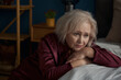 Sad elderly woman wearing pajamas sitting on bed feeling loneliness