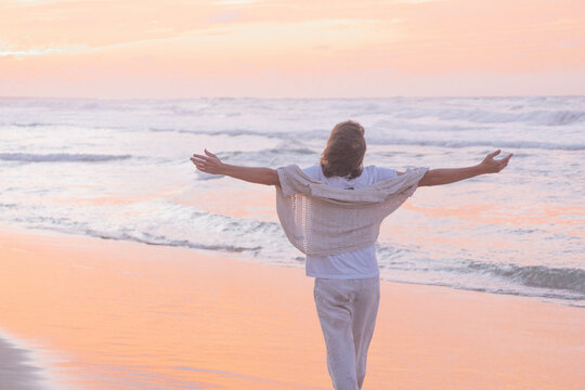 Tranquil scene of a person with outstretched arms facing the sea, bathed in the soft hues of a sunset on a peaceful, sandy shore