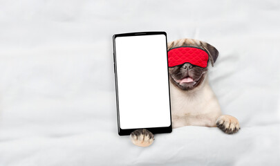 Fototapete - Pug puppy wearing sleeping mask lying on a bed at home and showing big smartphone with white blank screen in it paw. Top down view