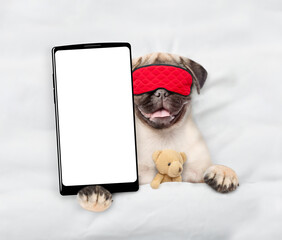 Canvas Print - Pug puppy wearing sleeping mask lying on a bed at home and showing big smartphone with white blank screen in it paw. Top down view