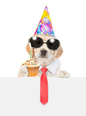 Wall Mural - Golden retriever puppy wearing sunglasses, necktie and party cap looks above empty white banner and shows cupcake with burning candle. isolated on white background