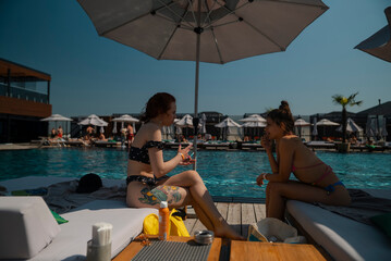 Wall Mural - Two young women in bathing suits sit together by the pool, chatting and laughing.