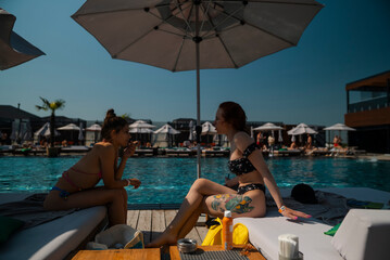 Wall Mural - Two young friends in swimsuits lounge by the poolside, enjoying each other's company.