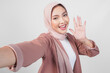 © Reezky - Cheerful young Asian Muslim woman in hijab posing for a selfie with waving hand hello isolated over white background.