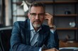 © marimalina - Photo of an attractive mature businessman sitting at his desk in the office. He is holding glasses and looking into the camera with confidence